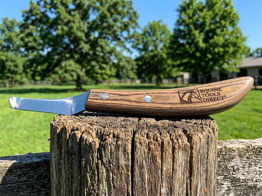 Equine Tools Direct curved hoof knife with natural wood 
handle resting on fence post in sunny outdoor farm setting